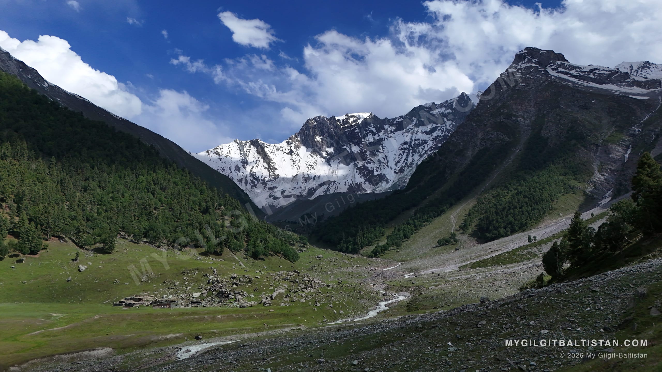 Stone summer huts on green meadow below a snowfield, where herders bring livestock during the warm months.